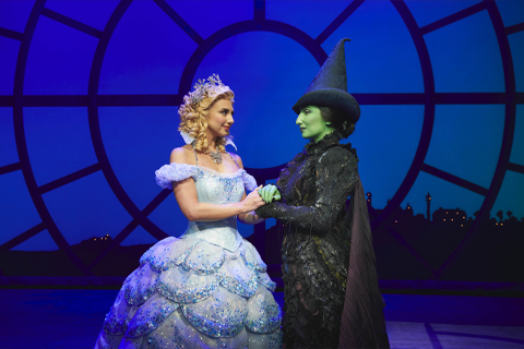 Glinda and Elphaba hold hands on stage beneath the Time Dragon clock